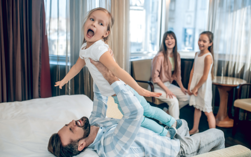 father holding child on bed and playing with mother and two girls playing in hotel room while dad is lifting energetic little girl up.