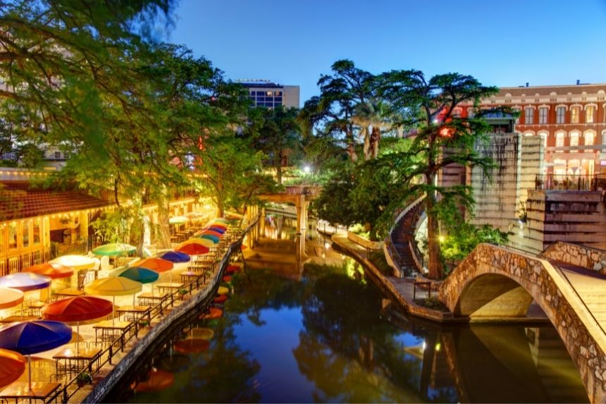 tables with colorful umbrellas, river, trees, and stone bridge on San Antonio River Walk