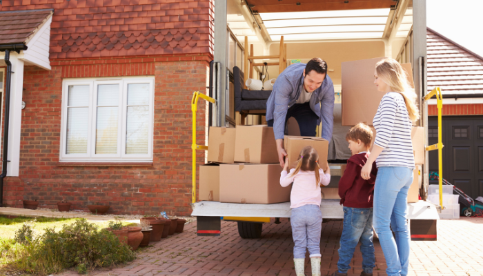 Dad bent over in moving truck in front of brick house to reach the cardboard box a little girl in pigtails is lifting up to him with mom in striped shirt and brother stand beside watching.