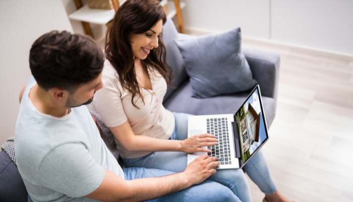 Man and woman sitting on gray couch smile at computer as they scroll real estate listing on computer sitting on their laps. 