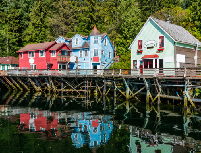 Photo of the historic boardwalk in Ketchikan, Alaska with vibrant colored buildings on dock with dark water below and a mature green trees behind.