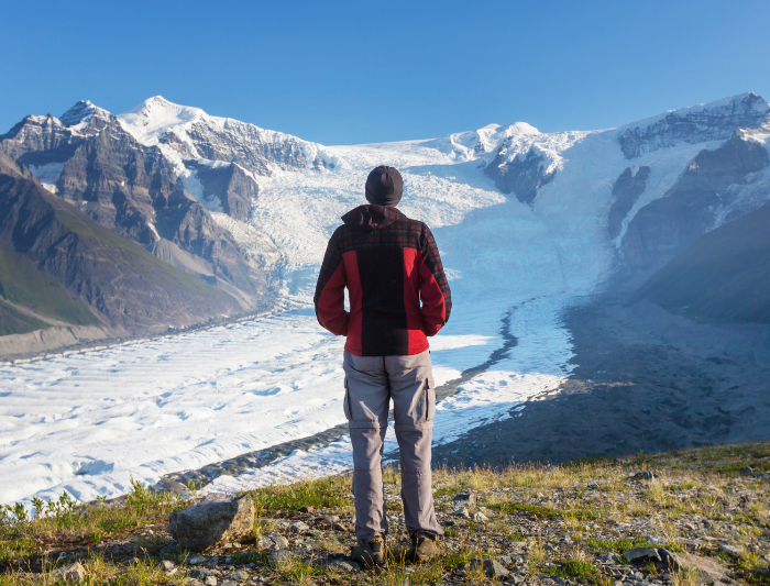 Hiker in red coat looks ahead at steep snow-covered path in Wrangell, St. Elias National Park, Alaska