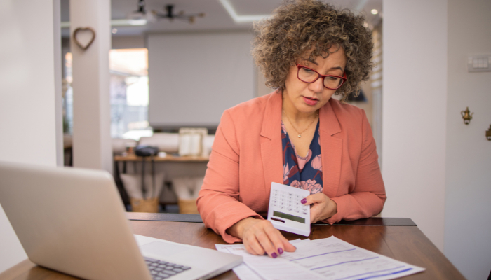 Woman wearing red glasses and orange blazer holds calculator as she looks at paperwork sitting in front of the computer.
