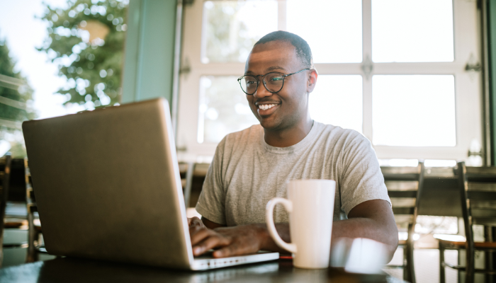 Man in glasses and gray t-shirt sits in bright room at table with white coffee mug and smiles while he types on computer in front of him.