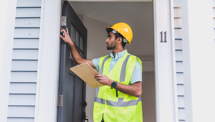 Man with short beard wearing a yellow hard hard and safety vest holding a clipboard inspection door jam of house.