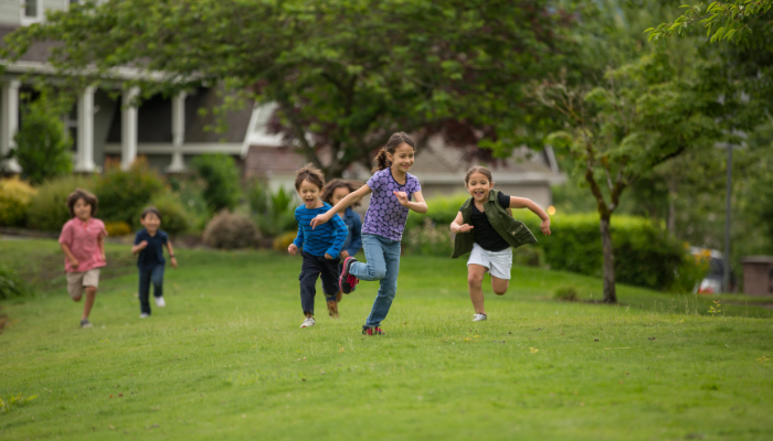 Group of kids running with smiles on their faces in green field with trees and houses behind them.