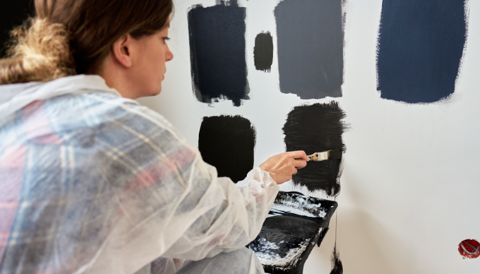 Woman with. hair pulled up in bun wears a smock over plaid shirt and paints test spots of varying dark colors on white wall.