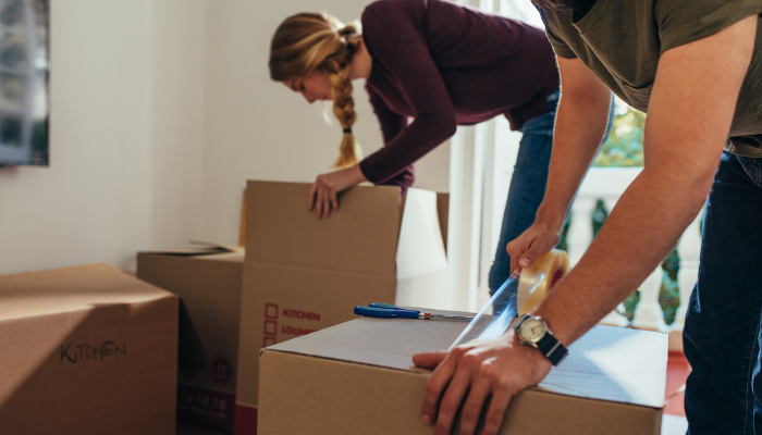 Man wearing t-shirt and watch tapes up moving box next to woman in purple long-sleeve shirt with braided hair packs up box.