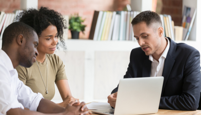 Man and woman wearing light green and white sit with hands folded on table listening to real estate professional in a suit coat talking while pointing to his laptop. 