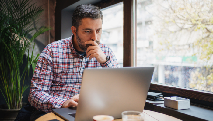 Man with beard wearing plaid long-sleeve shirt rests chin in hand while looking at computer on table in front of him. 