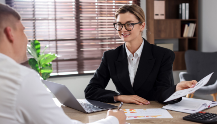 Woman in black blazer in white shirt and glasses smiles while holding paperwork up to man in white shirt sitting across the desk from her.