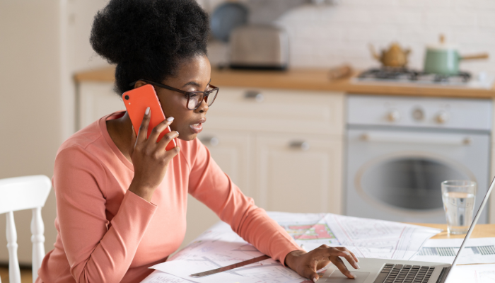 Woman in coral long sleeve shirt scrolls on computer while on a phone call on red phone. 