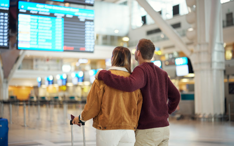 Man and woman stand with arms around each other with hand on suitcase looking at flight schedule in airport. 