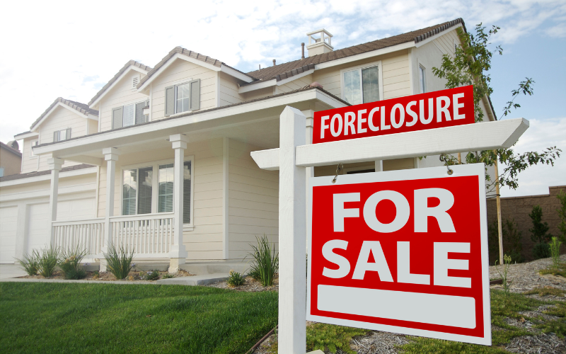 Photo of Foreclosure For Sale sign in front of beige house and green yard.