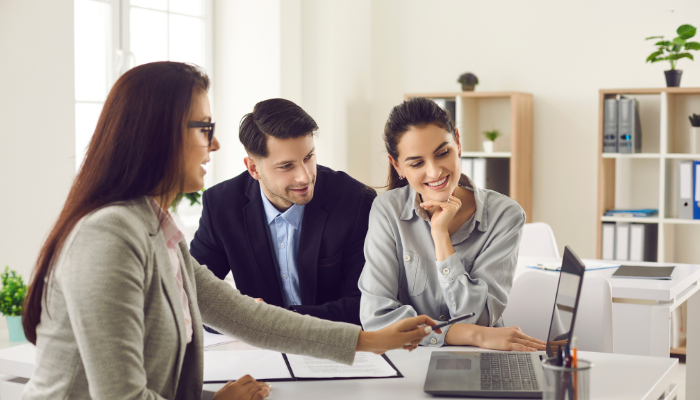 Man and woman meet with real estate agent while she points a pen at the computer in front of her.