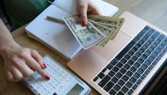 Woman with red nails budgeting in front of laptop with calculator and cash in hand.