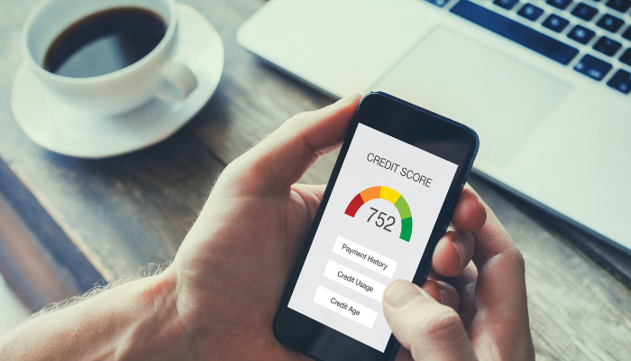 Man checking credit score on phone in front of computer and cup of coffee. 