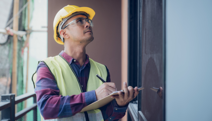 Home inspector in hardhat and vest taking notes on clipboard while inspecting house.