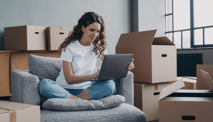 Woman with long, dark, curly hair wearing a white t-shirt and denim jeans sits cross-legged in chair holding computer in lap with cardboard boxes around the room.