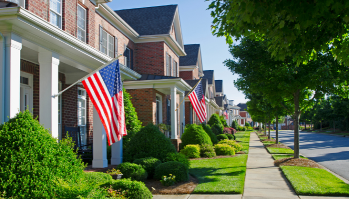 Quiet neighborhood road with brick homes featuring white columns and American flags hanging out front and lush green trees lining the road. 