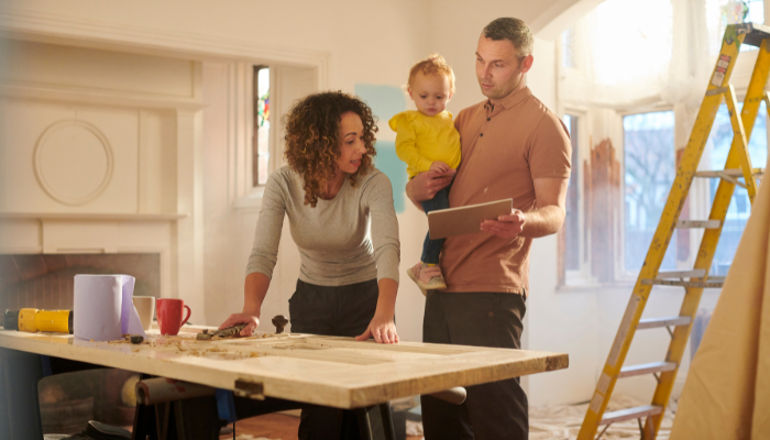 Man holding toddler in yellow shirt while showing woman with curly hair papers as they stand in room with ladder and renovation supplies.
