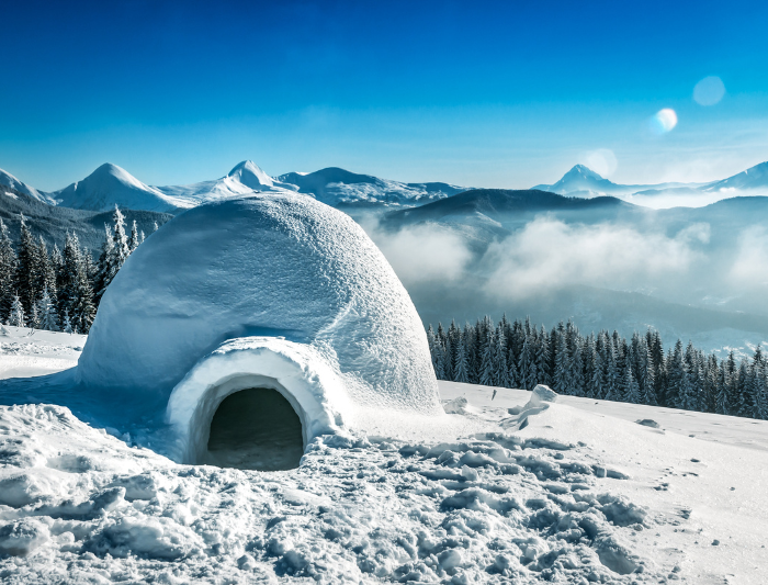 Large round igloo built on the side of a snowy mountain with a clear sky and sun shining above.