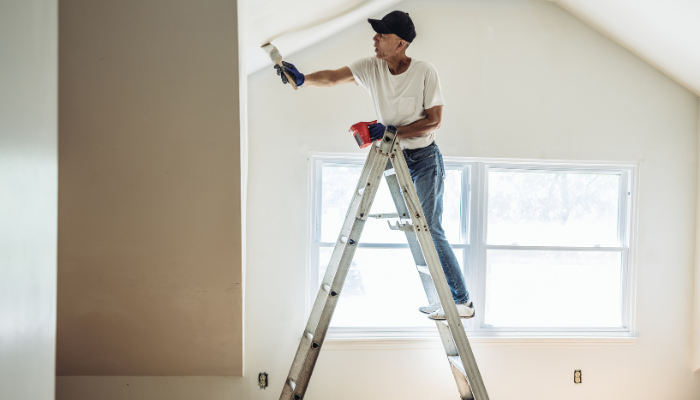 Man in black ball cap and white t-shirt stands on tall ladder in home to pain vaulted ceiling in home.