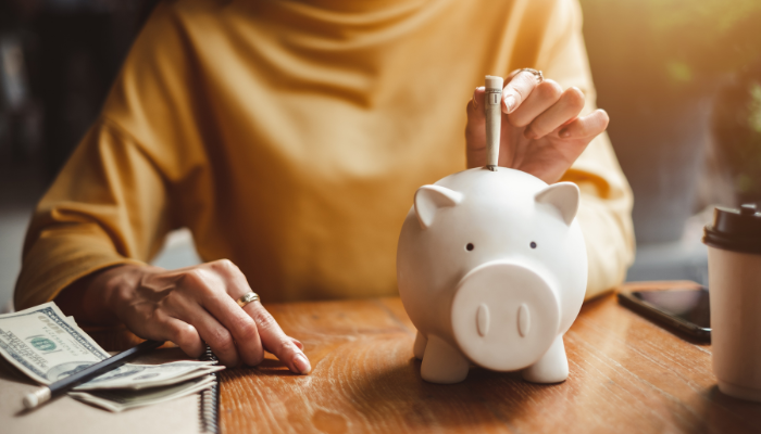 Woman in yellow turtle neck shirt sits at wooden table with a small stack of cash next to her and puts folded bill in white piggy back.