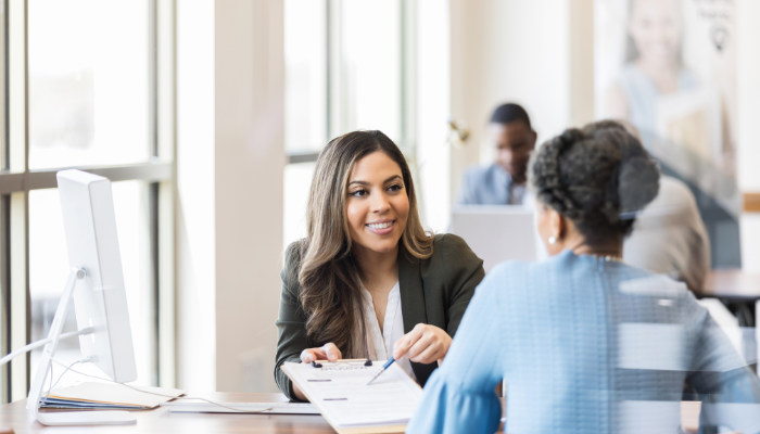 Loan officer in blazer uses pen to point to paperwork on clipboard as she smiles at woman in blue sitting across the desk from her.