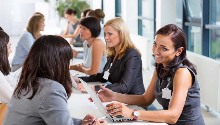 Two rows of women wearing neutral business attire sitting across a table from each other at a job fair. Women on the right side of the table have lanyards and computers.