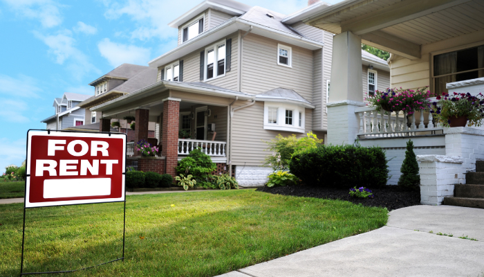 Red For Rent sign in green yard in front of homes with neutral siding, columns, and brick accents.