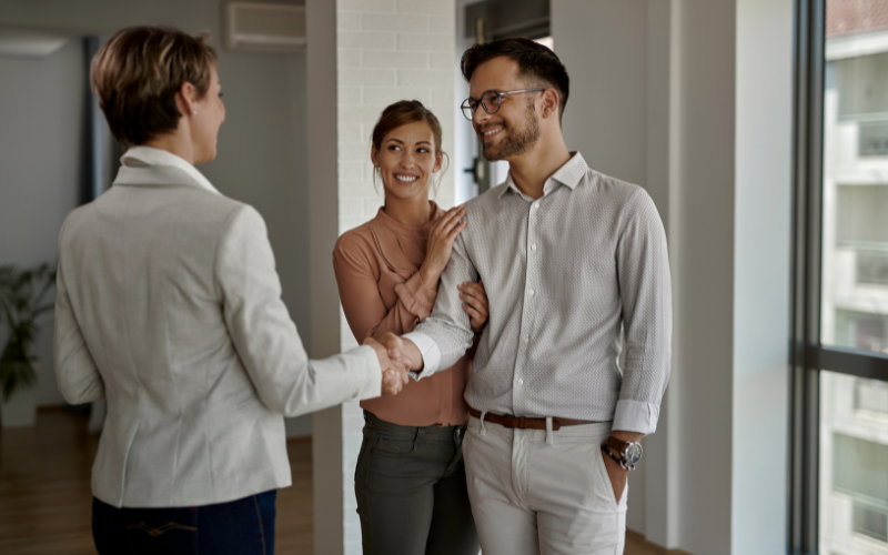Woman with hand on man smiling as he shakes hands with real estate agent in home. 