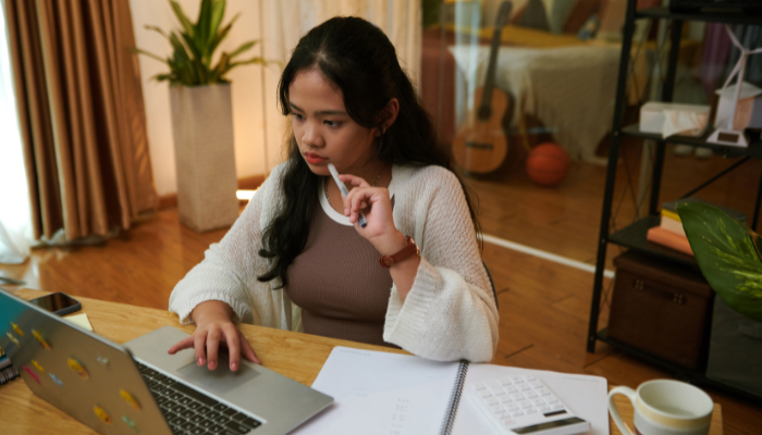 Girl wearing brown and tan concentrates on computer in front of her while she holds her pen to her mouth.
