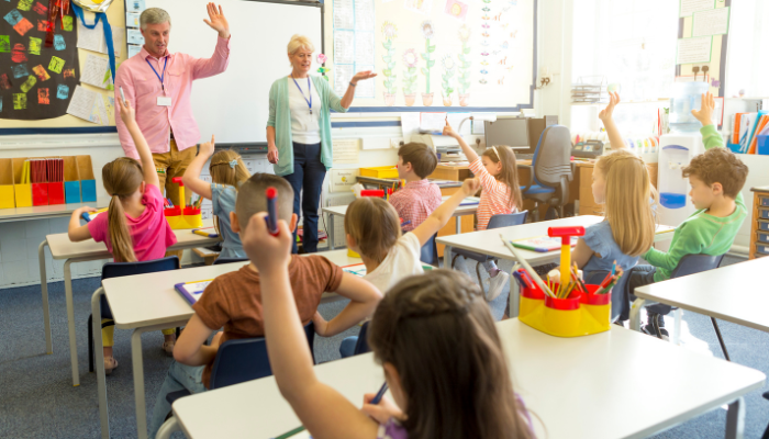 Colorful classroom with young kids and two teachers standing in the front wearing pink and green. 