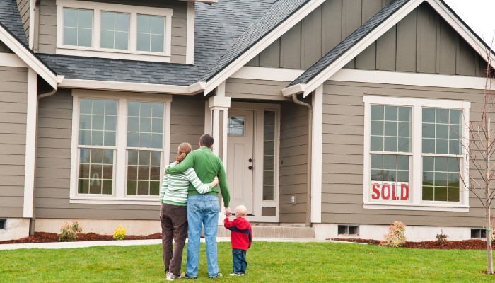 Family of three stand with their arms around each other facing beige home with sold sign in front of it.