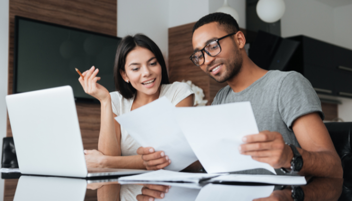 Couple wearing gray and white sit next to each other at table looking over paperwork in front of the computer.