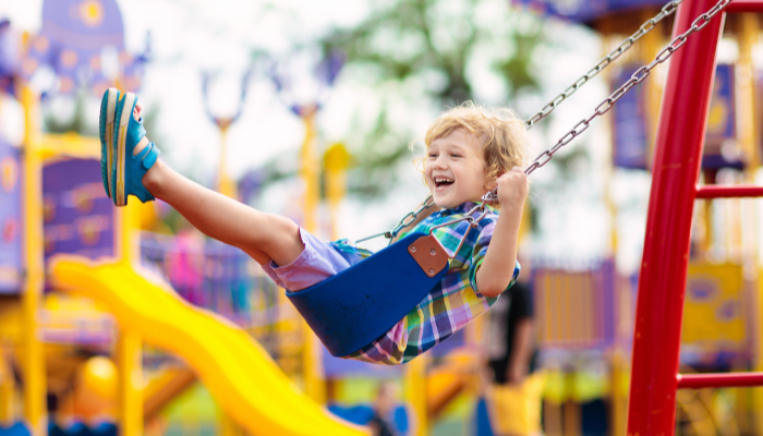Young boy in plaid shirt and blue shoes swings with smile on his face with a colorful playground structure in the background