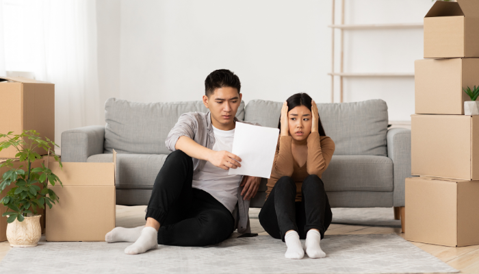 Distraught couple wearing neutral clothing sit on floor leaning on gray couch with moving boxes around them while looking at paperwork