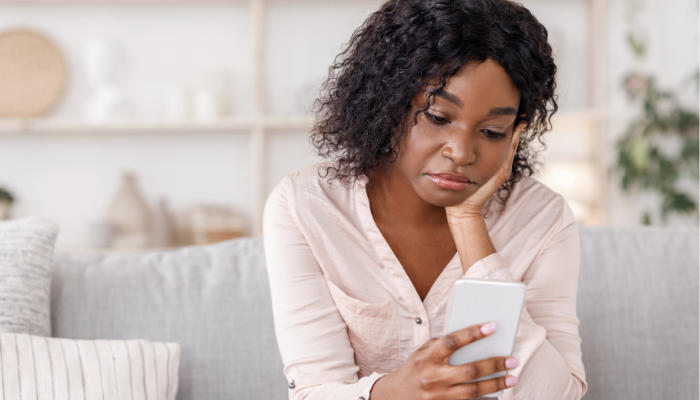 Woman in light pink shirt sits on light gray couch with head in hand looking concerned at her phone in hand.