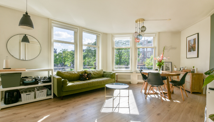 Airy and sunlit living room with green couch and dining chairs, light colored wooden floors.