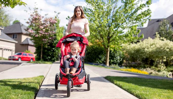 Mom in light pink shirt smiles and pushes red jogging stroller through the neighborhood with little girl sitting happy.