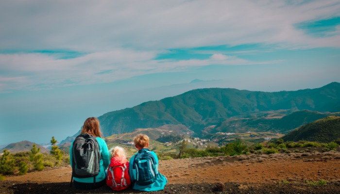 Mom and two kids sit in dirt with backpacks looking at mountains in Spain. 
