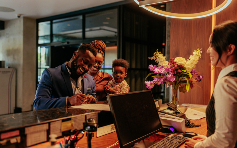 Man in blue suit with woman holding young child in arms check into beautiful hotel featuring warm lighting, a bouquet of flowers, and a smiling receptionist. 