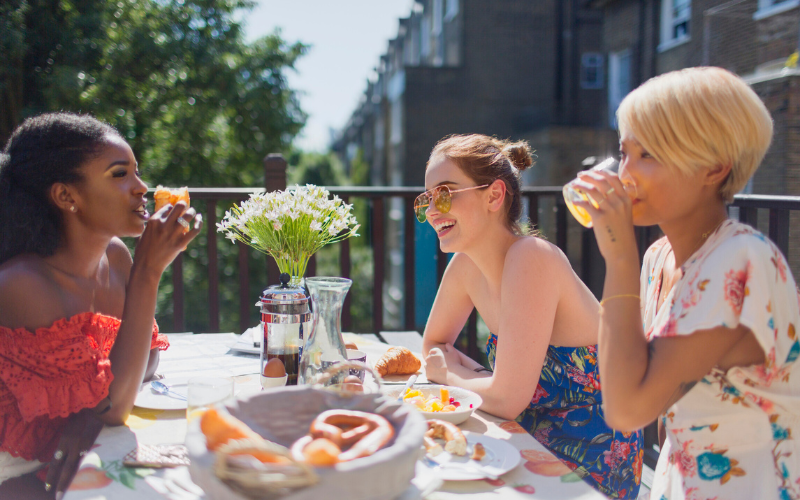 Three women sitting at an outdoor table with food, drinks, and a white tablecloth enjoying brunch together.