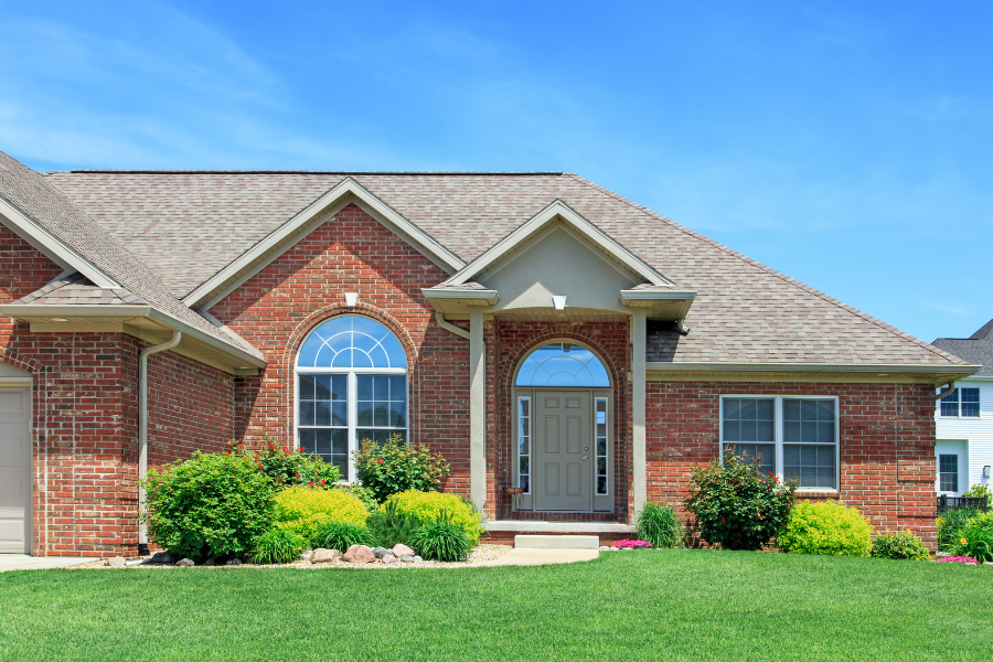 red brick one level home with shingled roof, green lawn and landscaping with blue sky in background
