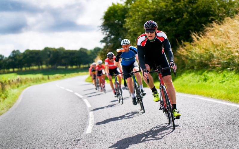 Cyclists on a group ride along marked, paved path, in park with greenery around. 