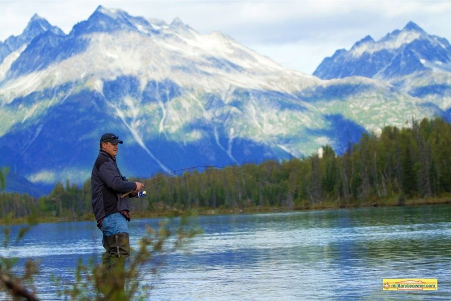 man fishing in waders in river with snow covered mountains in background in Alaska
