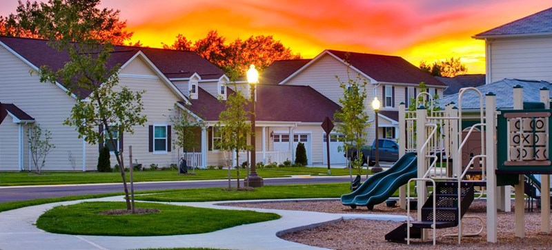 Bolling on base military family housing. There is a sunset in the background. The street lights are on. The homes are a mix of single and two-story. The are light tan with white trim. In the foreground is a park with a playset.