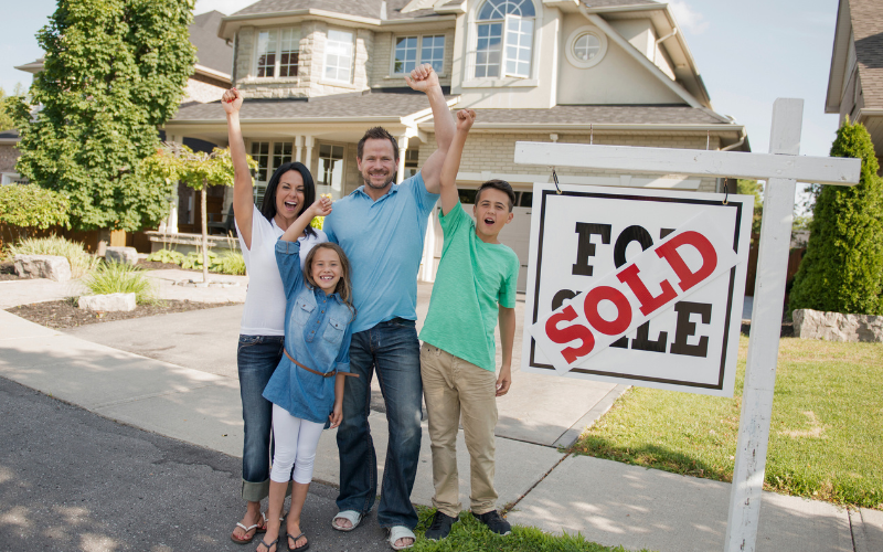 Family of four wearing, white, blue, and green stand with one arm raised cheering in front of Sold sign outside beige home with grow trees in the yard.