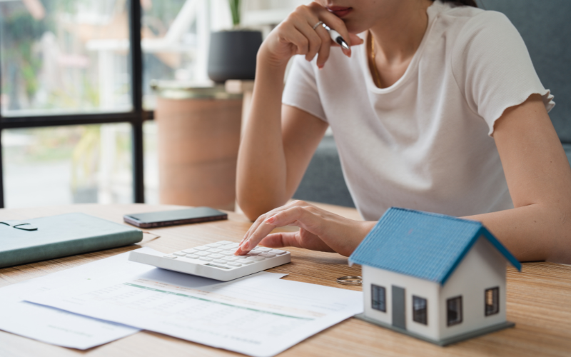 Woman at desk thinking about buying a house with calculator, paperwork, and model house next to her. 